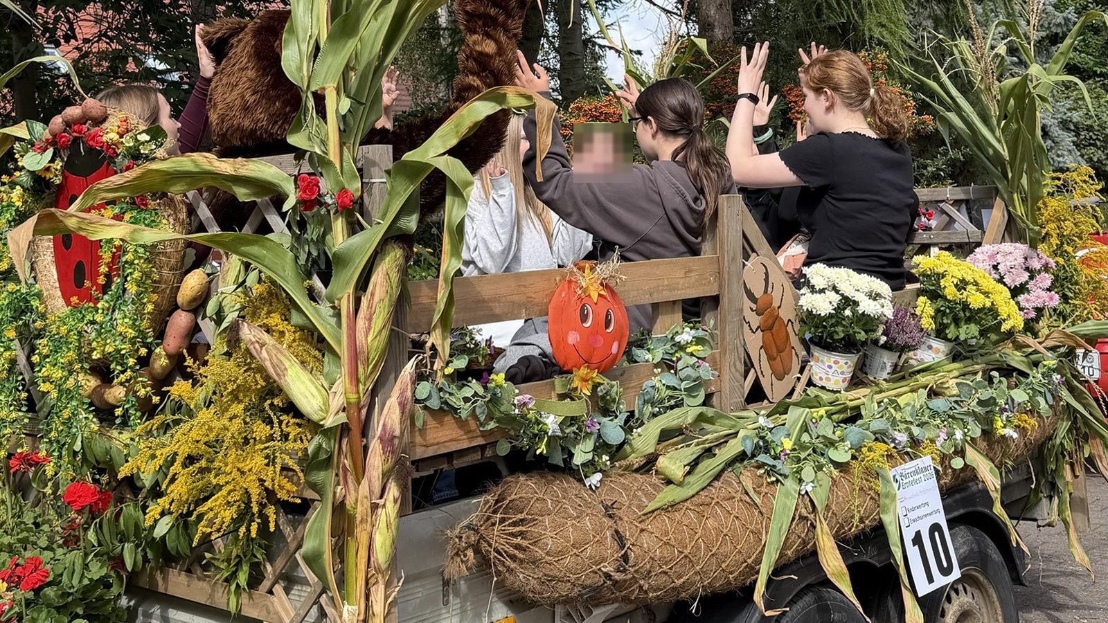 12 von 12 September 2026 - mit Maisstauden, Blumen und einem lachenden Kürbis geschmückter Erntewagen, darauf winkende Jugendliche