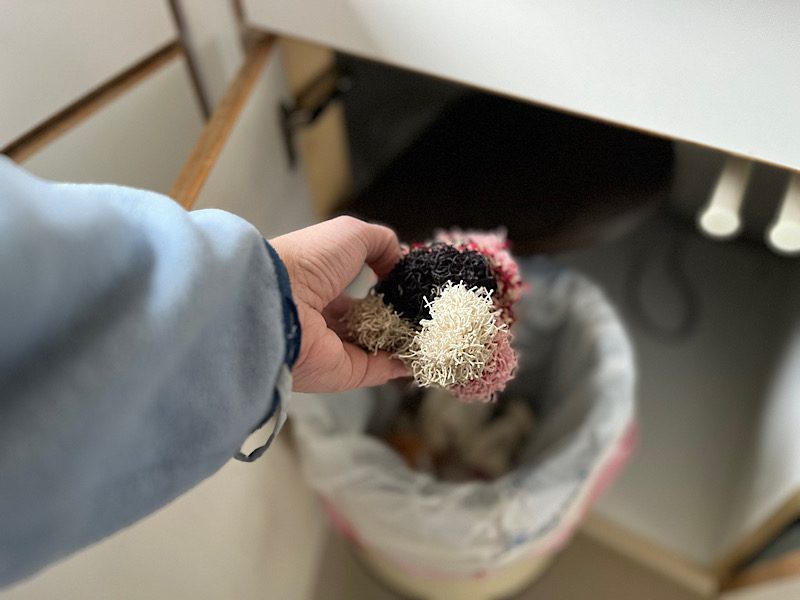 Hand hält drei bunte Spülschwämme aus Garn über einen Stoffbeutel unter der Spüle