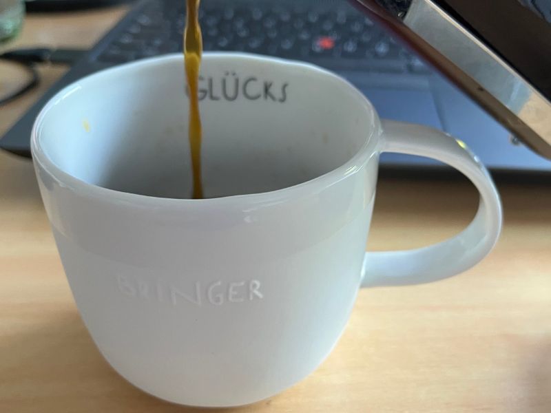 12 von 12 - August 2025 - 3: weiße Glücksbringer-Tasse vor Laptop, in die Kaffee gegossen wird.