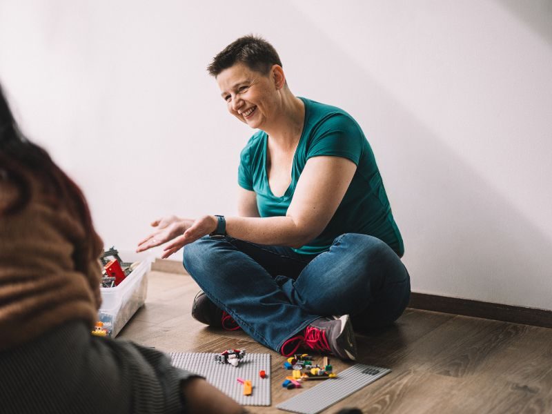 Kindercoachin Danielle Berg sitzt lachend am Boden und arbeitet in einer Coaching-Sitzung mit einer Kiste LEGO-Steine.