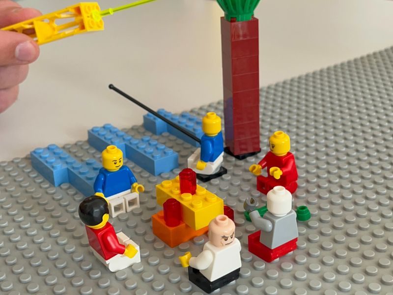 Mehrere LEGO-Minifiguren auf einer Bauplatte, eine Hand greift mit einem Bauteil in die Szene – eine Situation wird mit LEGO nachgebaut.