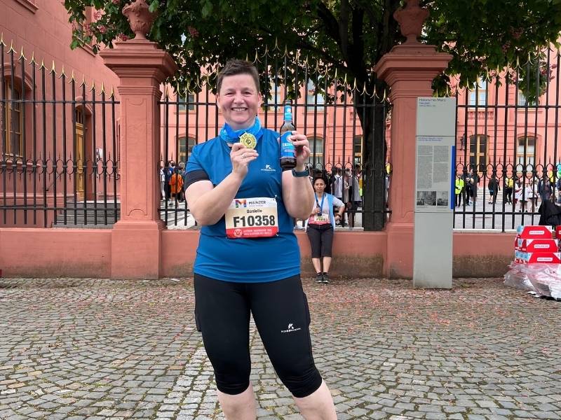 Danielle Berg vorm Mainzer Schloss mit Startnummer, Medaille und Finisher-Bier beim Mainzer Gutenberglauf