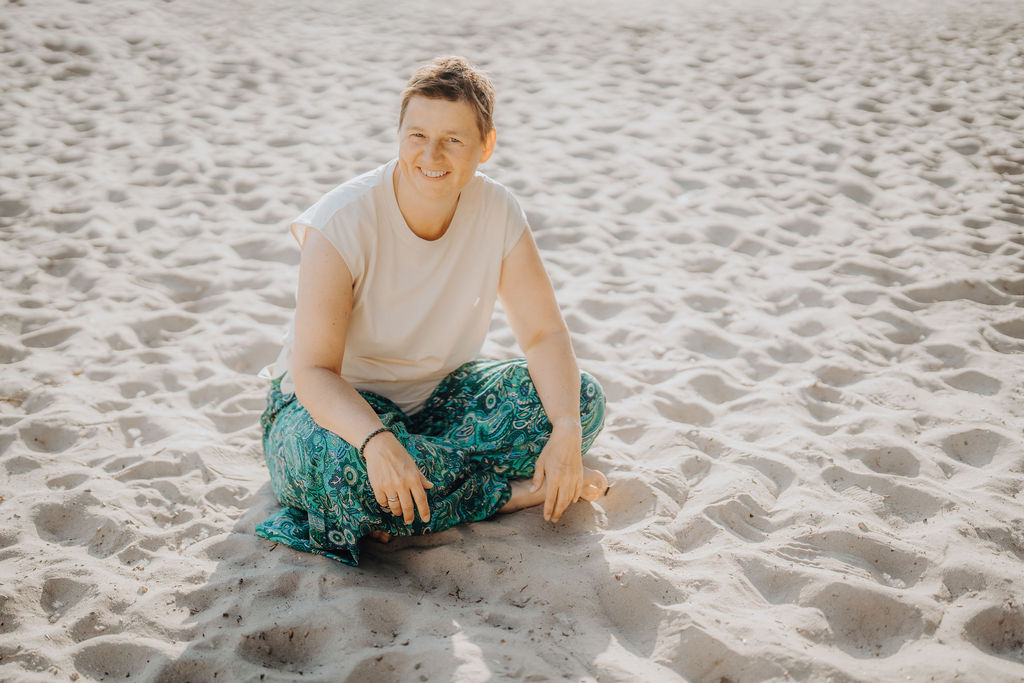 Danielle Berg sitzt barfuß im Schneidersitz im Sand, trägt ein helles Shirt und eine gemusterte petrolfarbene Hose und lächelt in die Kamera.