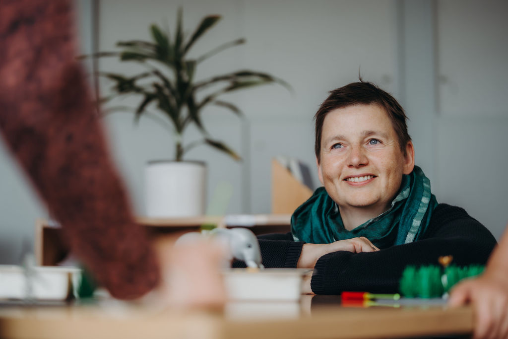 Danielle Berg sitzt an einem Holztisch mit LEGO® Steinen und einer kleinen Elefantenfigur und lächelt zu jemandem außerhalb des Bildes.